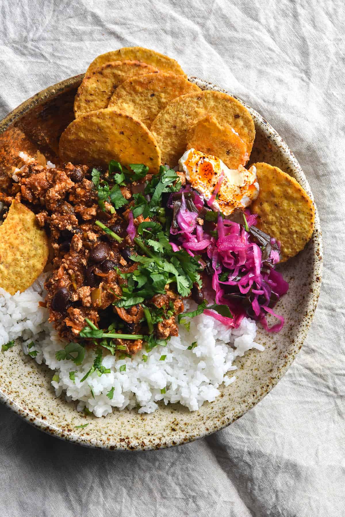 An aerial close up view of a bowl of colourful FODMAP friendly vegetarian chilli. The chilli is in a beige speckled open faced ceramic bowl which sits atop a beige linen backdrop. In the bowl is rice, chilli, pickled red cabbage, corn chips, lactose free sour cream topped with chilli oil and coriander. The bowl is casually arranged