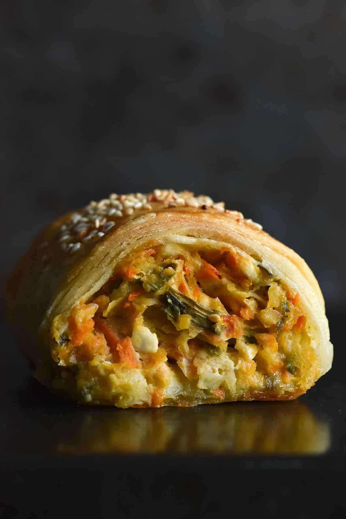 A side on close up macro image of a gluten free vegetable, feta and chickpea sausage roll. The roll sits atop a dark grey background against a dark backdrop. The side on angle reveals the vegetable and melty cheese filling inside, as well as the flaky, golden pastry encompassing it. The roll is topped with toasted sesame seeds that are visible atop the roll