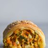 A side on close up macro image of a gluten free vegetable, feta and chickpea sausage roll. The roll sits atop a white marble table against a white wall. The side on angle reveals the vegetable and melty cheese filling inside, as well as the flaky, golden pastry encompassing it. The roll is topped with toasted sesame seeds that are visible from the angle
