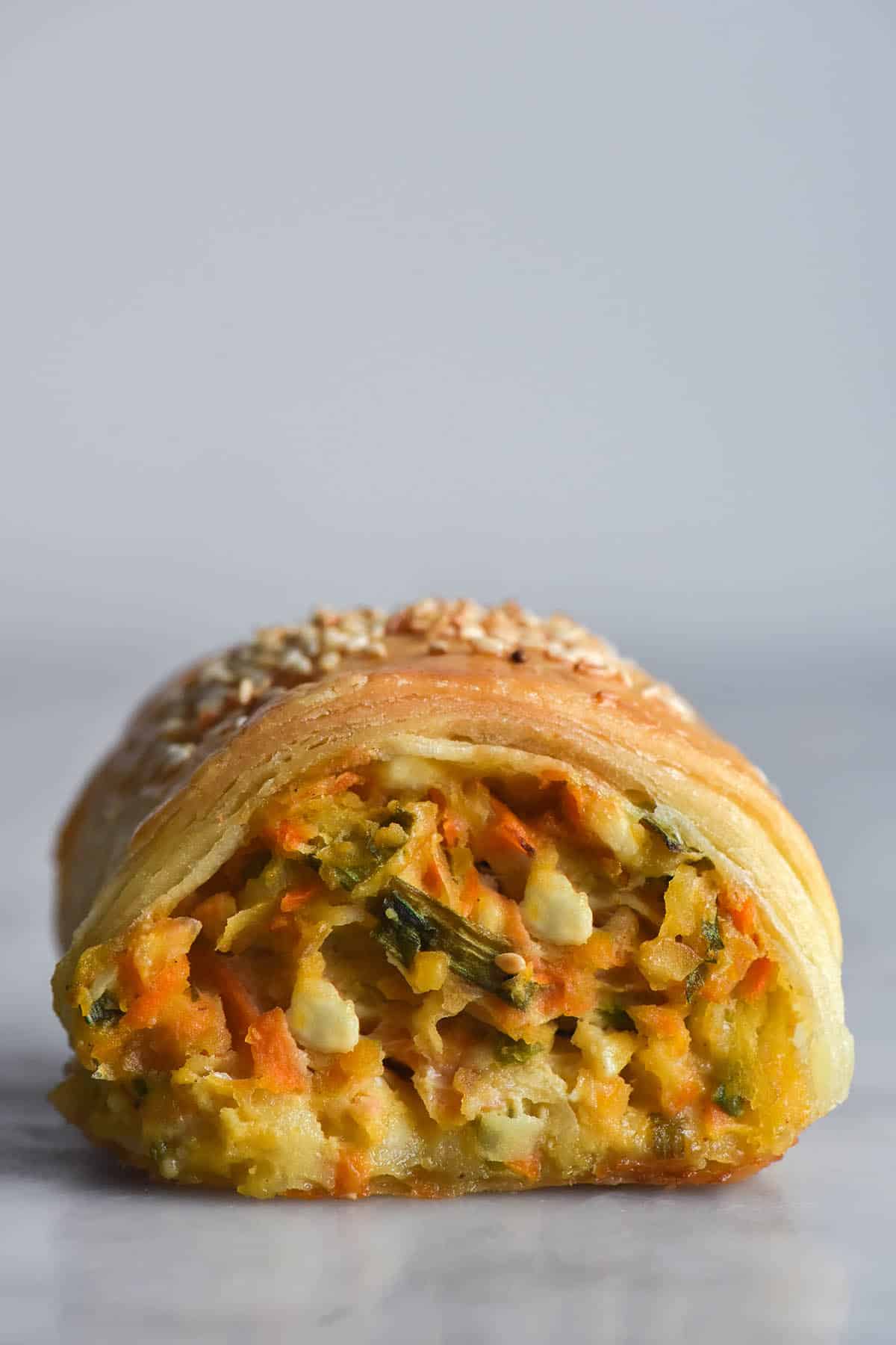 A side on close up macro image of a gluten free vegetable, feta and chickpea sausage roll. The roll sits atop a white marble table against a white wall. The side on angle reveals the vegetable and melty cheese filling inside, as well as the flaky, golden pastry encompassing it. The roll is topped with toasted sesame seeds that are visible from the angle