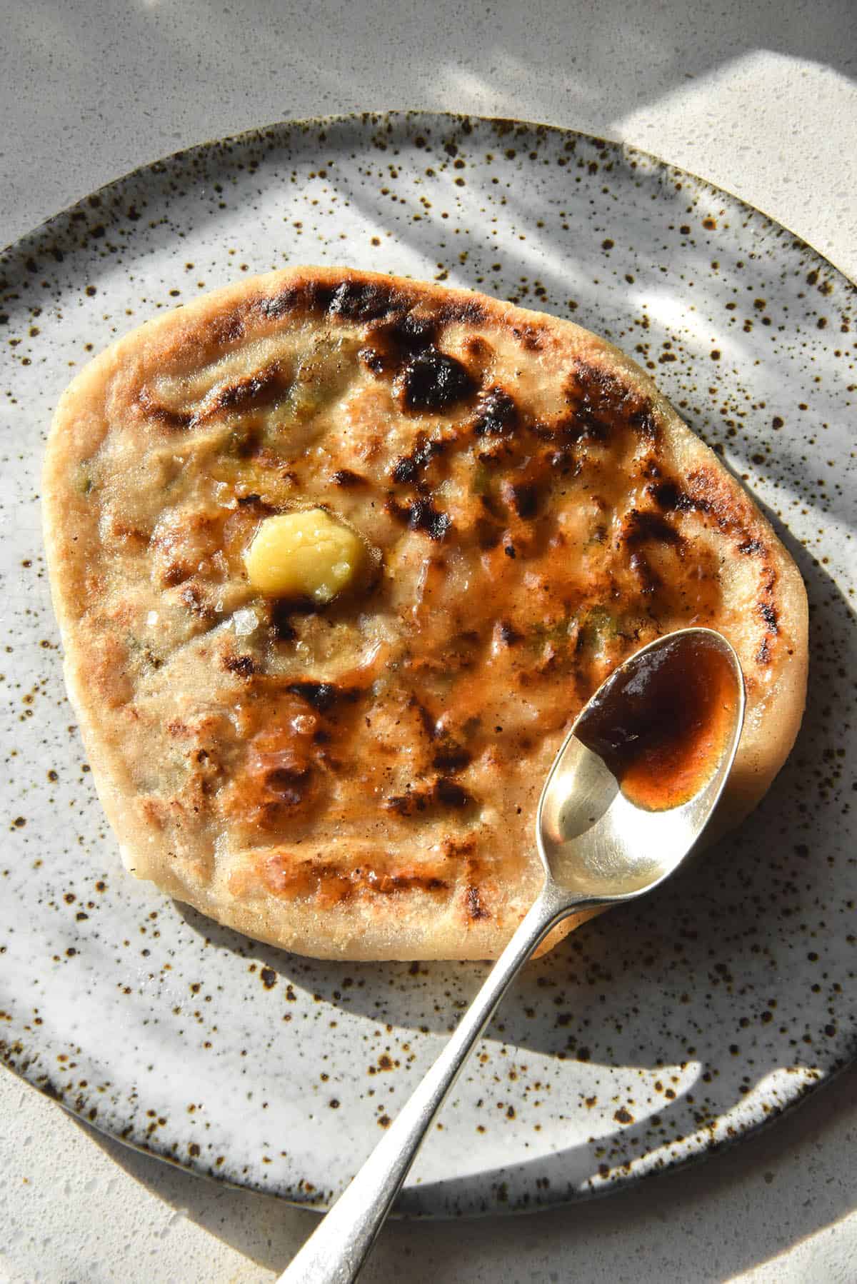 Gluten free Aloo Paratha topped with imli chutney and garlic infused ghee with sea salt flakes. The paratha sits atop a white speckled ceramic plate in bright sunlight atop a white countertop. A spoonful of imli chutney sits on the bottom right half of the paratha. 