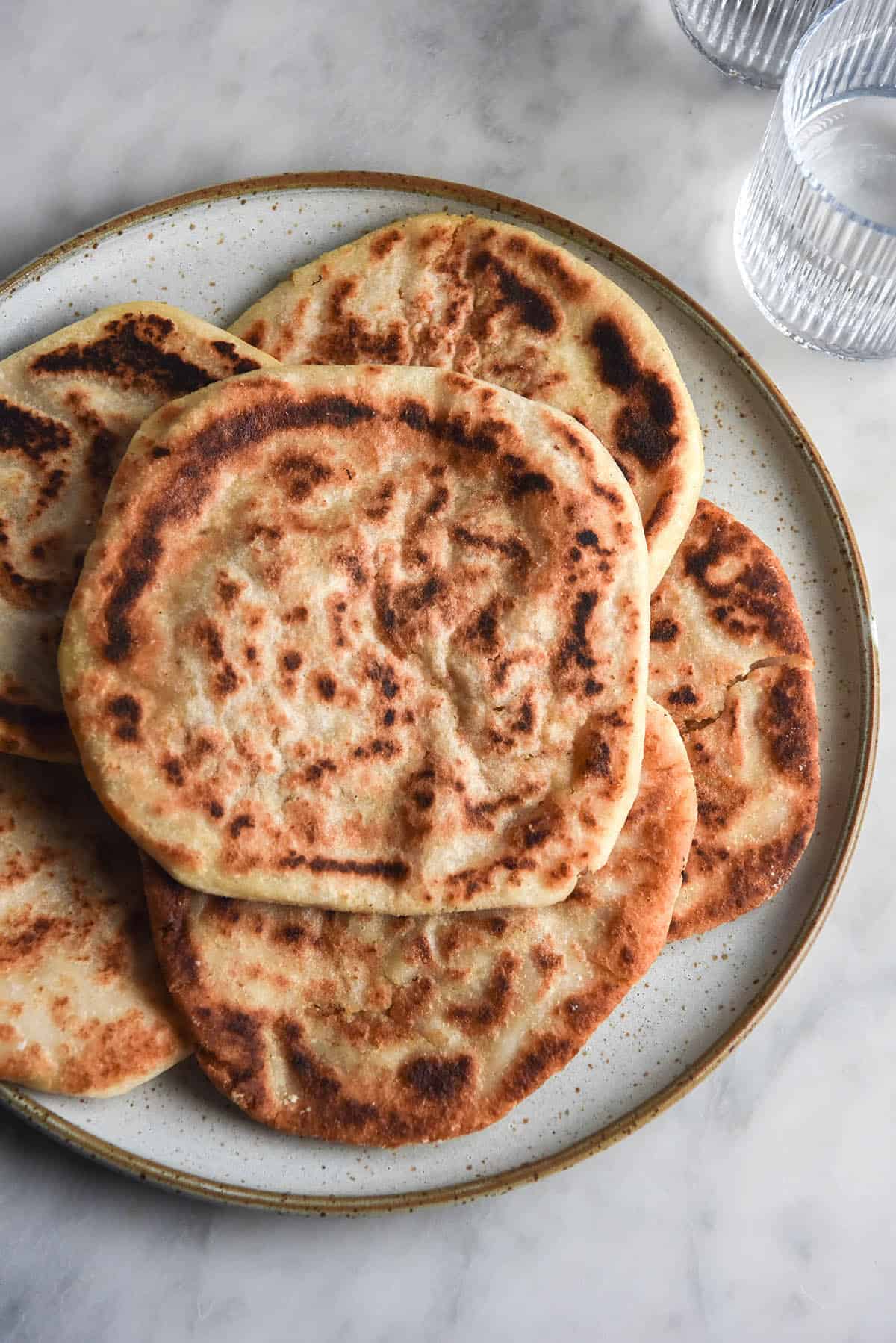 An aerial view of a white ceramic plate stacked with gluten free aloo parathas. The parathas are speckled with golden brown frying marks and the plate sits atop a white marble table with water glasses in the top right corner. 