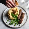A photo of a white speckled ceramic plate topped with vegan sausages, mash and peas. A hand extends out across the white marble table to pour gravy over the mash potato and some sunlit glasses of water sit to the top right of the image