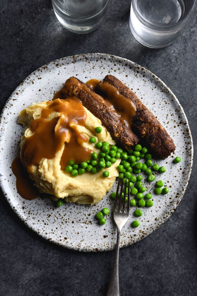 An aerial view of a plate of vegan and FODMAP friendly bangers and mash with peas and gravy. The food is served on a white ceramic plate on a dark blue backdrop.