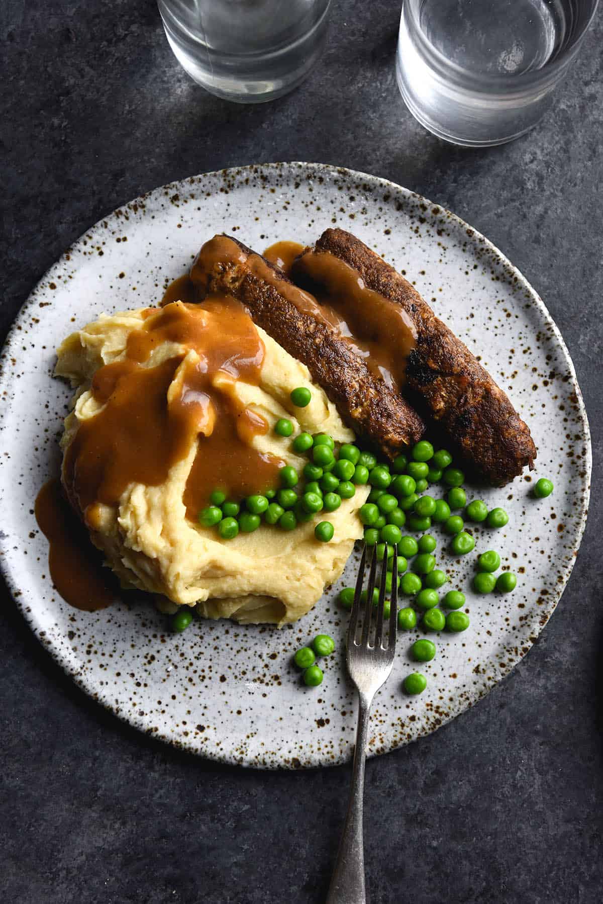 An aerial view of a white speckled ceramic plate topped with gravy covered mash, peas and vegan sausages. The plate sits atop a dark blue textured backdrop and a fork extends off the plate to the bottom right of the image. 