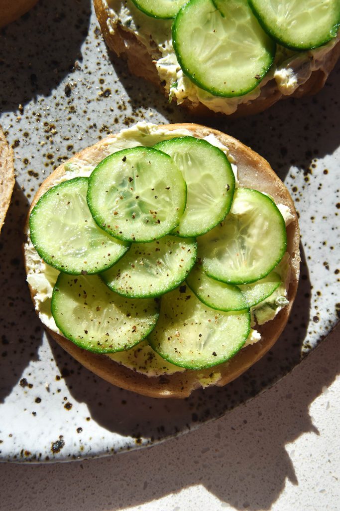 An aerial view of gluten free bread rolls, halved and topped with whipped goat's cheese and cucumber slices. The bread roll halves sit atop a white speckled ceramic plate on a white countertop. They are bathed in natural sunlight, creating shadows to the bottom right of the image.