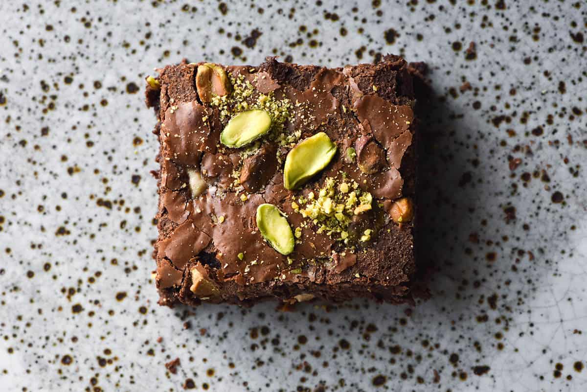 An aerial macro photo of a gluten free brownie square atop a white speckled ceramic plate. The brownie has a shiny, flaked top that is covered with sliced pistachios with a smattering of bright green pistachio dust. 