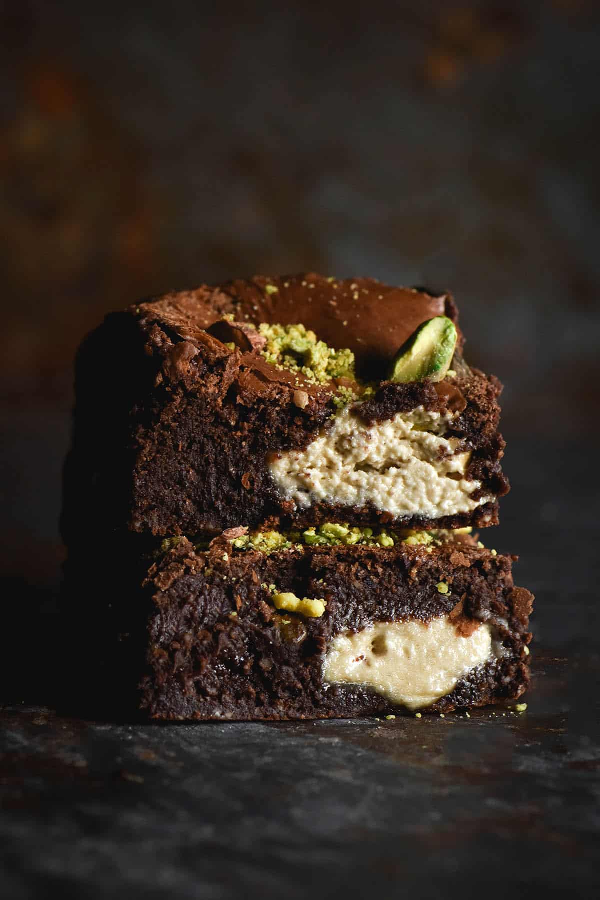 A side on view of a stack of two gluten free brownie squares that are filled with halva and pistachios. Large chunks of halva are visible in the brownie sides, as are chunks of browned and fresh, bright green pistachios. The brownies sit atop a dark blue metal surface and in front of a dark grey rusted backdrop