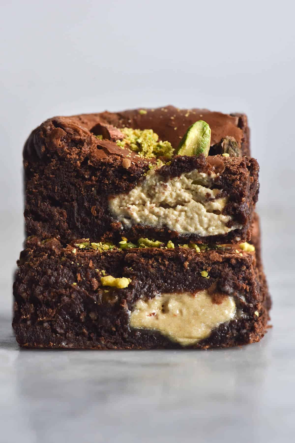 A side on view of a stack of two gluten free brownie squares that are filled with halva and pistachios. Large chunks of halva are visible in the brownie sides, as are chunks of browned and fresh, bright green pistachios. The brownies sit atop a white marble table against a white backdrop