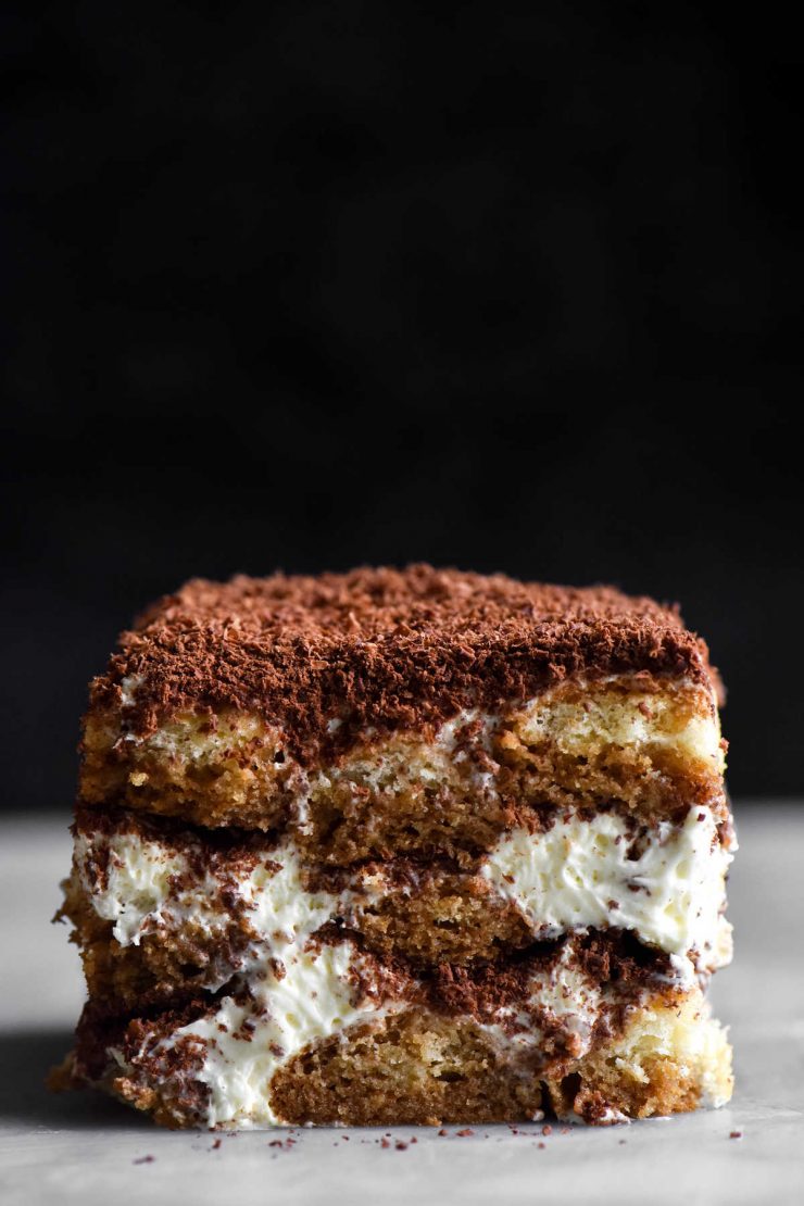 A close up, side on view of a slice of gluten free, FODMAP friendly tiramisu. The slice sits atop a marble table which contrasts with a black background