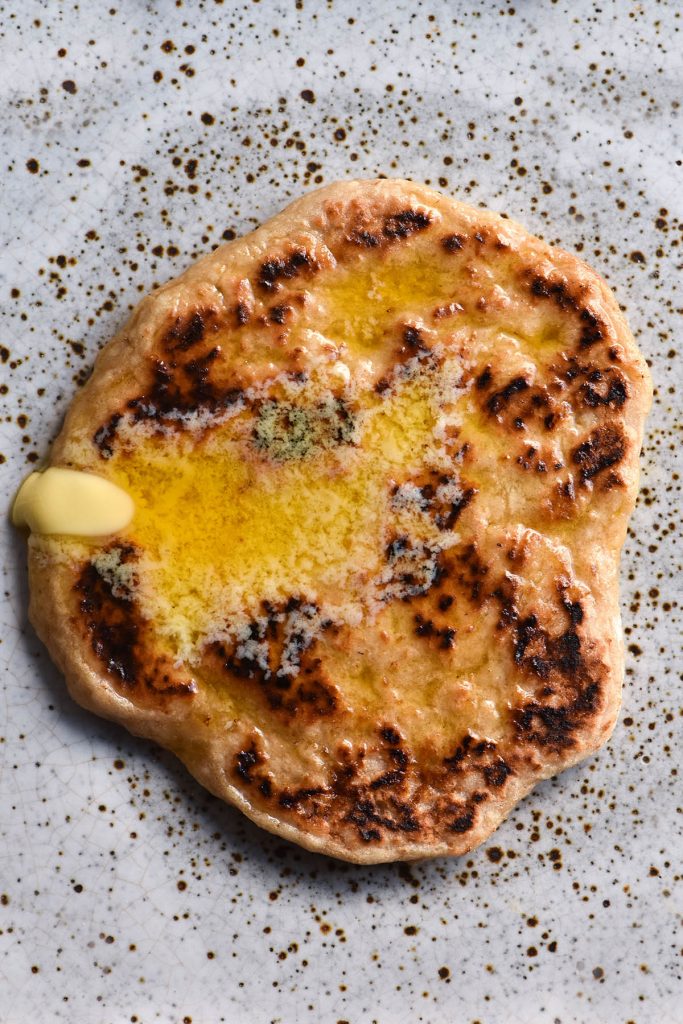 An aerial close up view of a gluten free yeasted flatbread topped with melted butter and sitting atop a white speckled ceramic plate