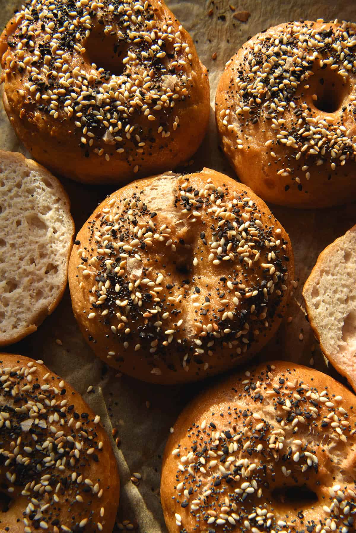 A close up aerial image of gluten free bagels topped with everything bagel seasoning, sitting atop a lined baking tray. The bagels sit in warm sunlight. 
