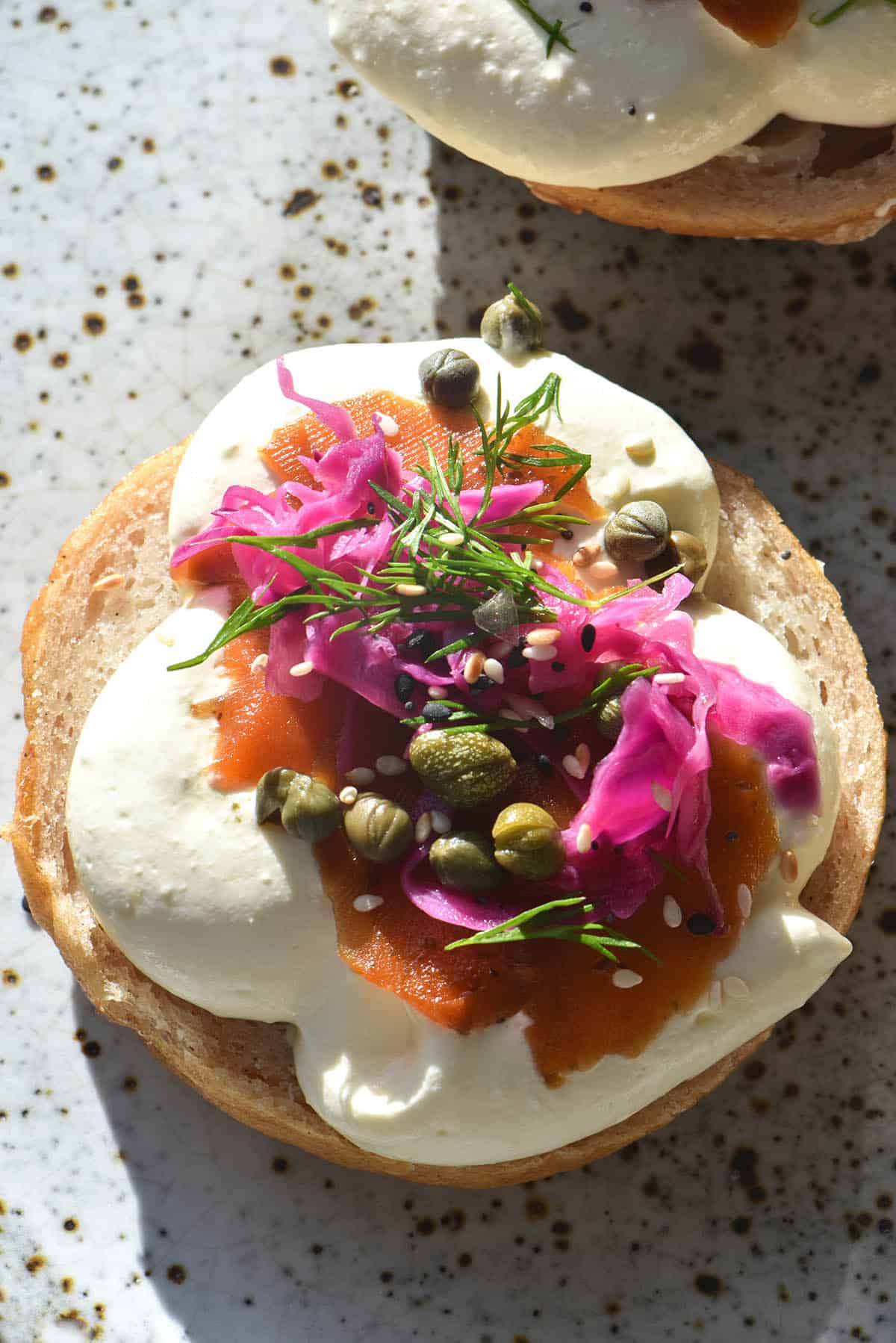 an aerial view of a gluten free bagel half topped with FODMAP friendly 'pickled onion, easy carrot lox, capers and whipped mascarpone. The bagel sits atop a white ceramic plate bathed in sunlight. 