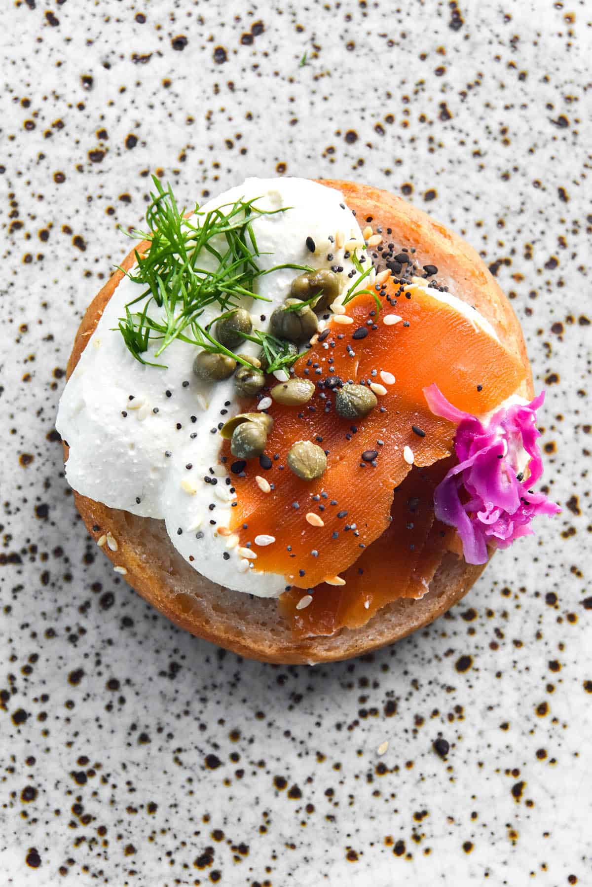 An aerial image of a gluten free bagel half topped with whipped mascarpone cream, carrot lox, capers, dill, FODMAP friendly 'pickled red onion' and FODMAP friendly everything bagel seasoning. The bagel sits in the centre of a white speckled ceramic plate