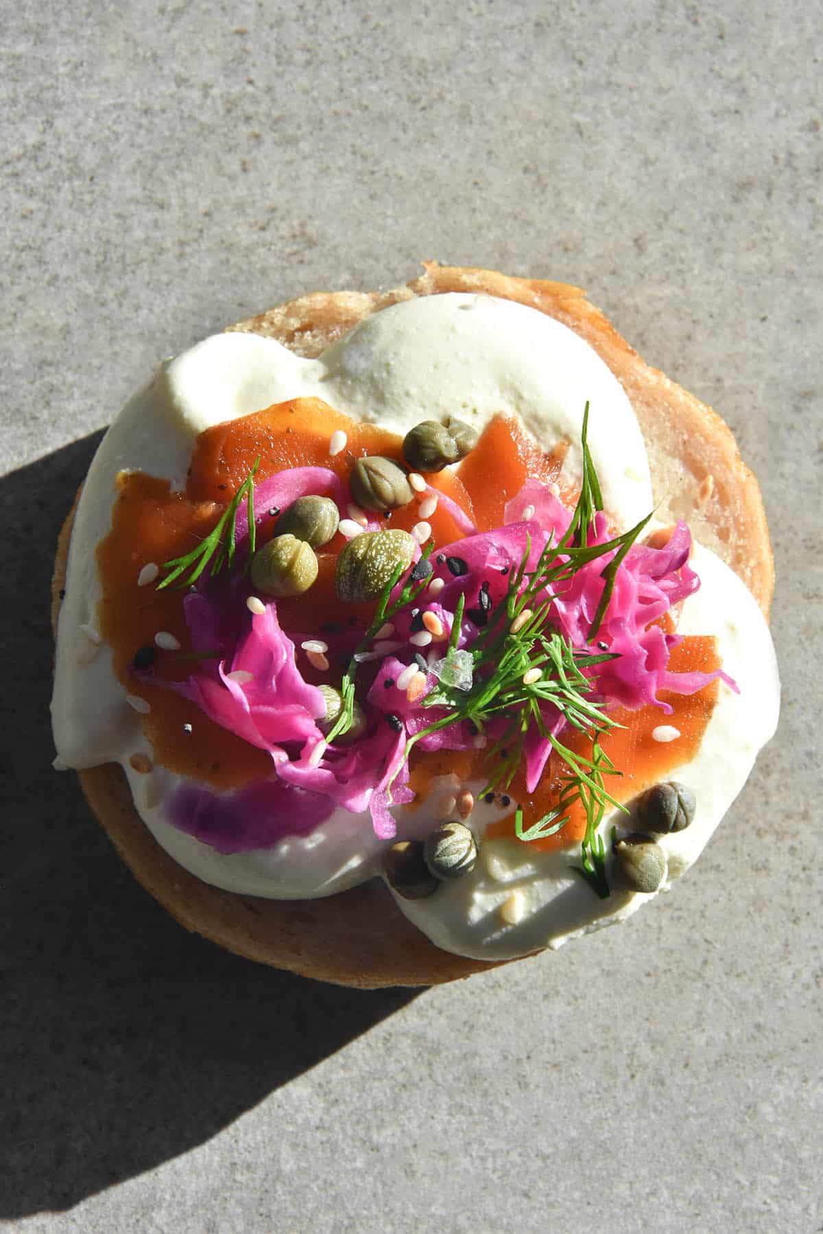An aerial image of a topped bagel half bathed in bright sunlight on a grey stone background. The bagel is topped with whipped lactose free mascarpone, easy carrot lox, FODMAP friendly pickled onions and capers.