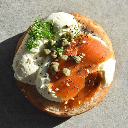 An aerial view of a gluten free bagel half topped with whipped mascarpone cream, carrot lox, capers, dill and FODMAP friendly everything bagel seasoning. The bagel sits atop a grey stone backdrop in harsh sunlight, and the shadow extends to the bottom right hand side of the image