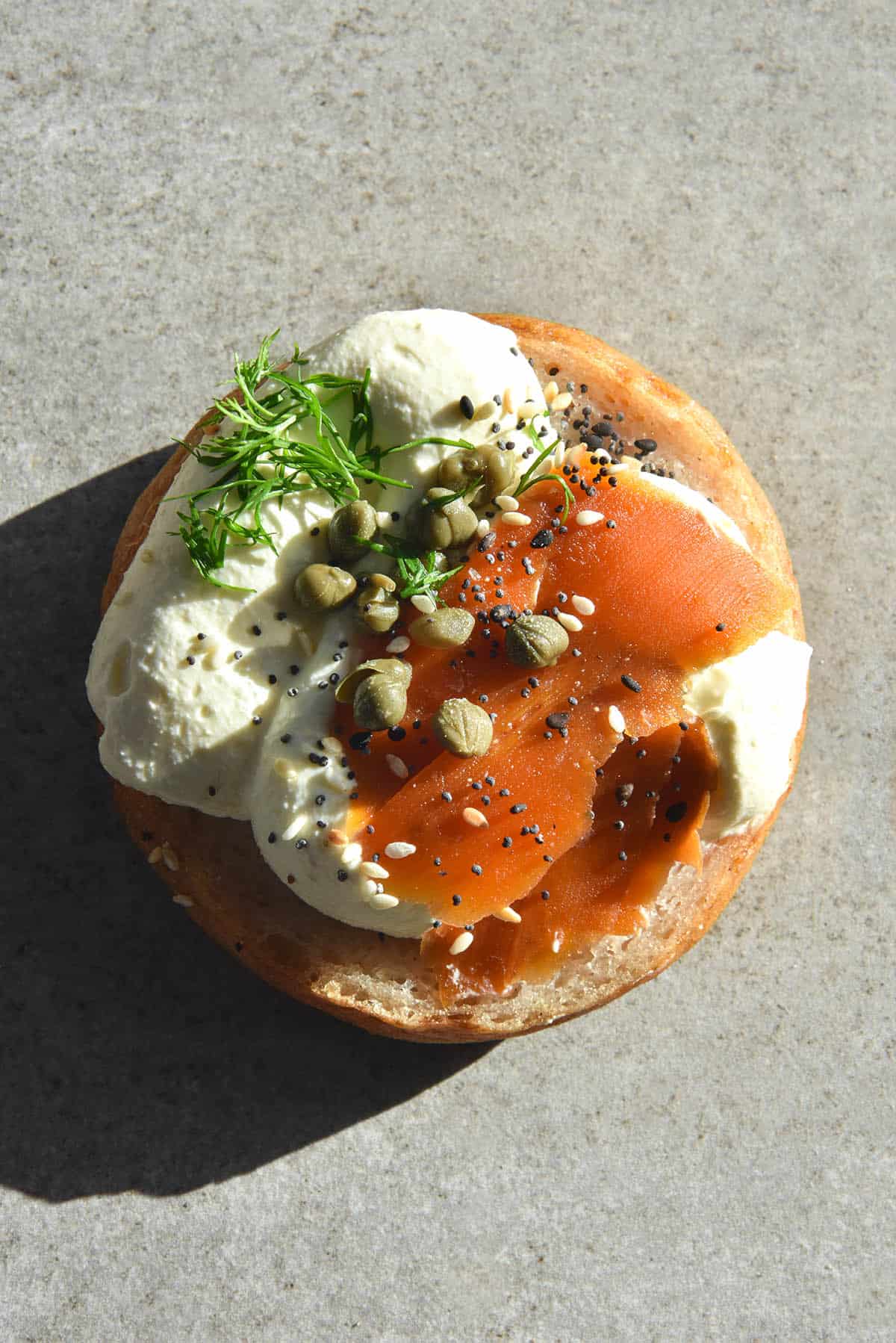 An aerial close up image of a gluten free bagel topped with lactose free mascarpone, carrot lox, capers, dill and a sprinkle of FODMAP friendly everything bagel seasoning. It sits atop a light grey stone backdrop in harsh sunlight, and the bagel's shadow extends to the bottom right of the image