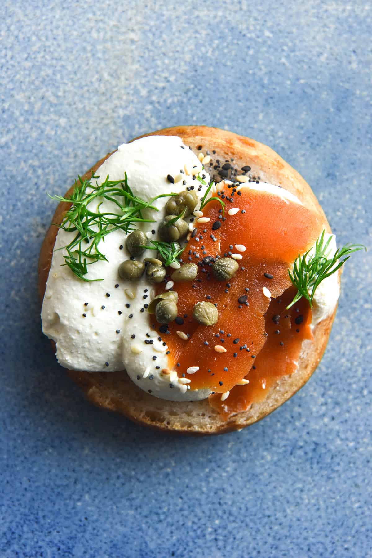 An aerial view of a gluten free bagel half atop a sky blue ceramic plate. The bagel is topped with whipped lactose free mascarpone, easy carrot lox, capers and dill, along with a sprinkle of FODMAP friendly everything bagel seasoning.