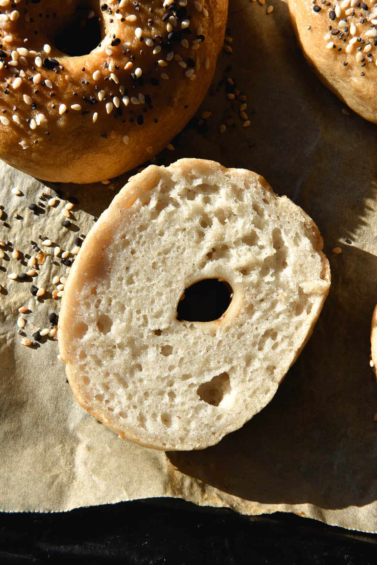 An aerial close up view of the crumb of a gluten free bagel. The bagel has been sliced and sits crumb side up on a lined baking sheet. It has an open crumb and is surrounded by other bagels and sprinkles of everything bagel seasoning 