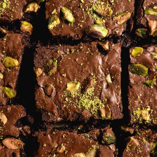 An aerial close up photo of a sliced slab of halva and pistachio chocolate brownies. The top of the brownies is shiny and dotted with halva pieces, pistachios and extra pistachio dust.