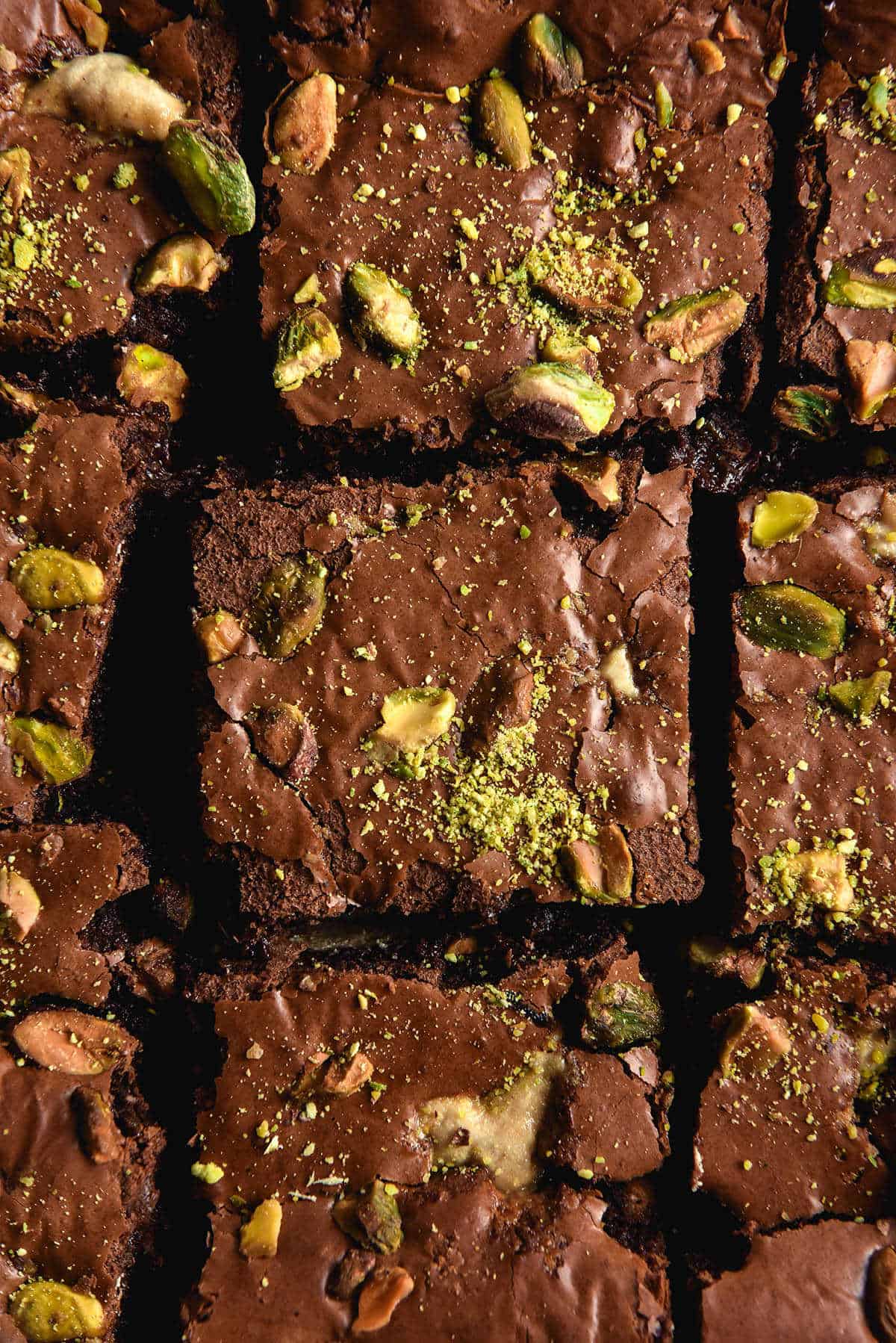 An aerial close up photo of a sliced slab of halva and pistachio chocolate brownies. The top of the brownies is shiny and dotted with halva pieces, pistachios and extra pistachio dust.