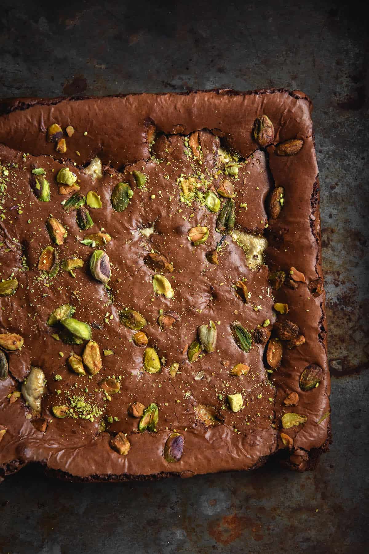 An aerial macro view of a slab of gluten free chocolate brownie filled with pieces of halva and topped with chopped pistachios. The slab sits atop a dark metal rusty backdrop.