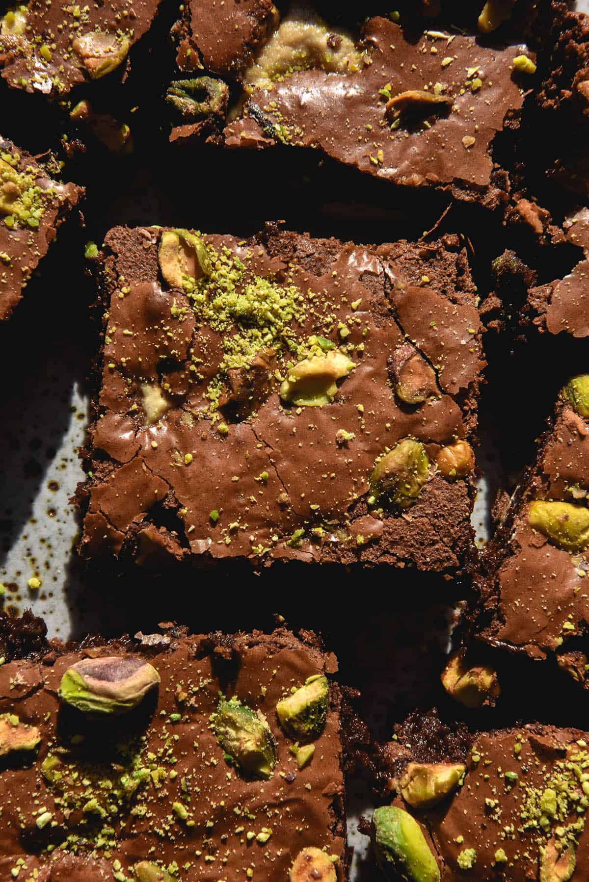 An aerial, macro photo of a white speckled ceramic plate topped with gluten free pistachio and halva brownie squares. The brownie tops are crackled and shiny and have pistachios, pieces of halva and extra bright green pistachio dust sprinkled on top. 