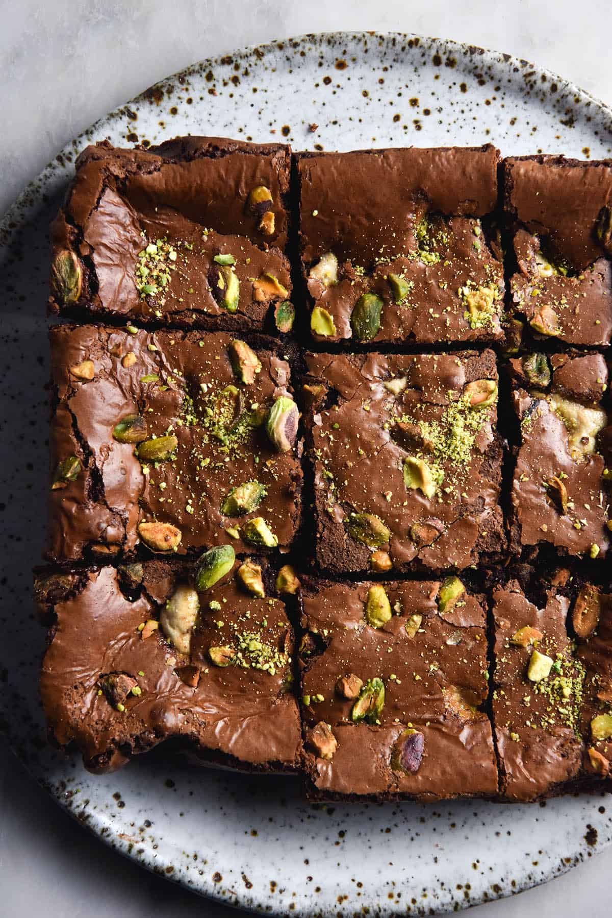 An aerial close up photo of a sliced slab of halva and pistachio chocolate brownies. The top of the brownies is shiny and dotted with halva pieces, pistachios and extra pistachio dust. The slab sits atop a white speckled ceramic plate on a white marble backdrop