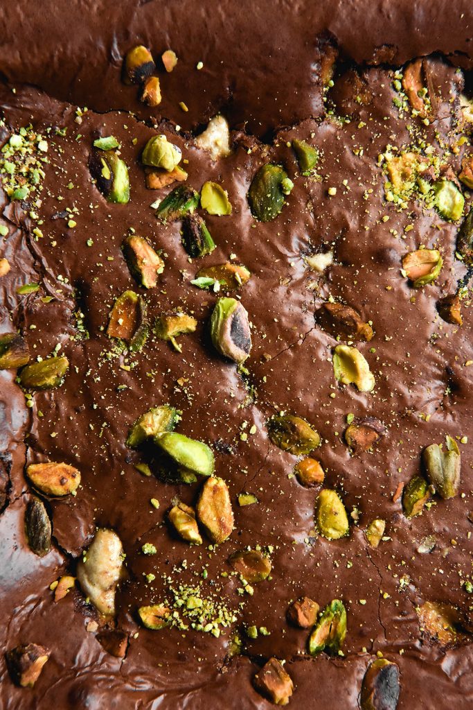 An aerial macro photo of a slab of gluten free brownies topped with pistachios and halva. The pistachios contrast against the shiny, crackled top of the brownie with their bright green colour