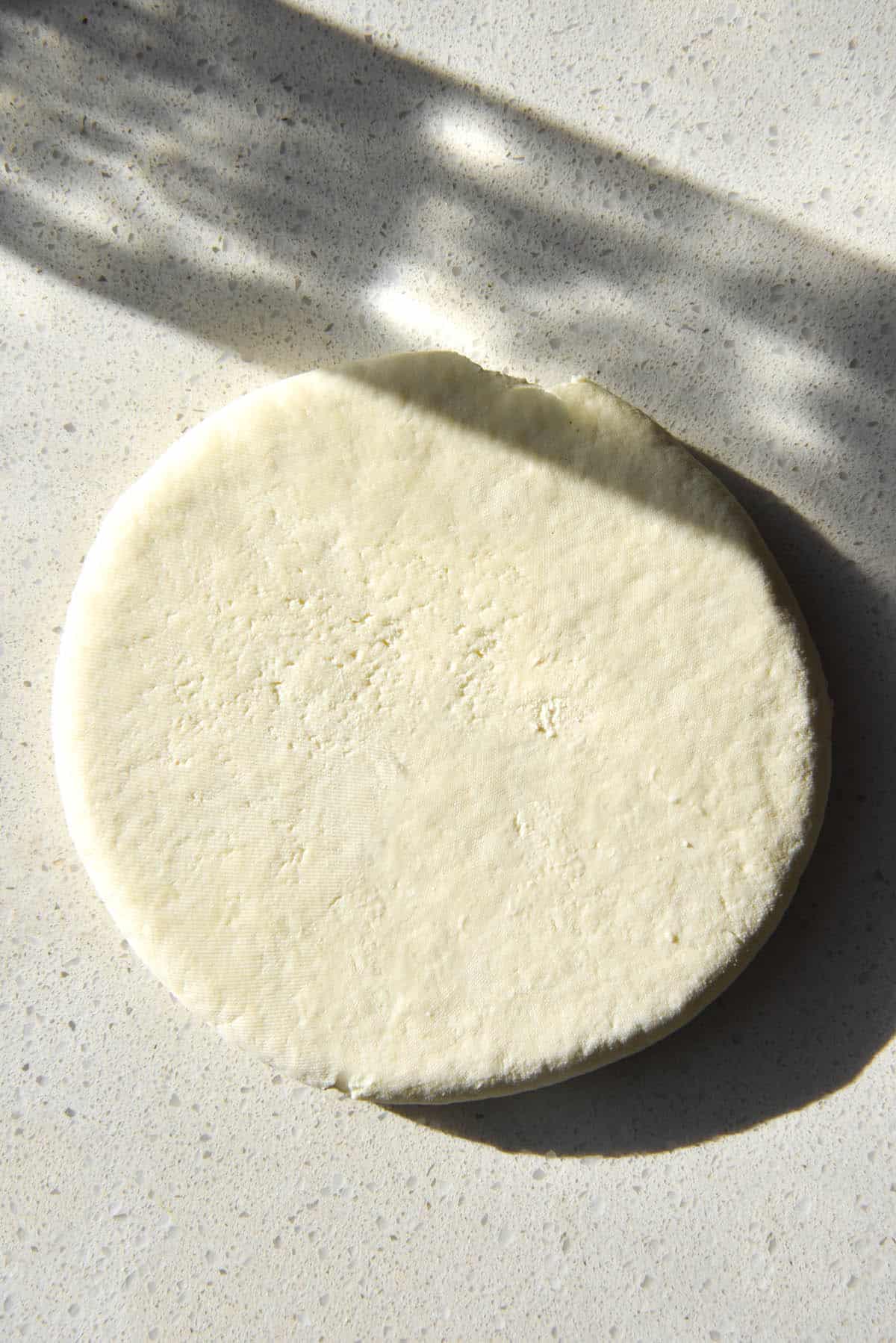 An aerial image of a disk of homemade lactose free paneer on a white kitchen counter. The disk of paneer sits in the centre of the image and a shadow of a water glass casts a pattern over the top of the paneer disk and the counter.