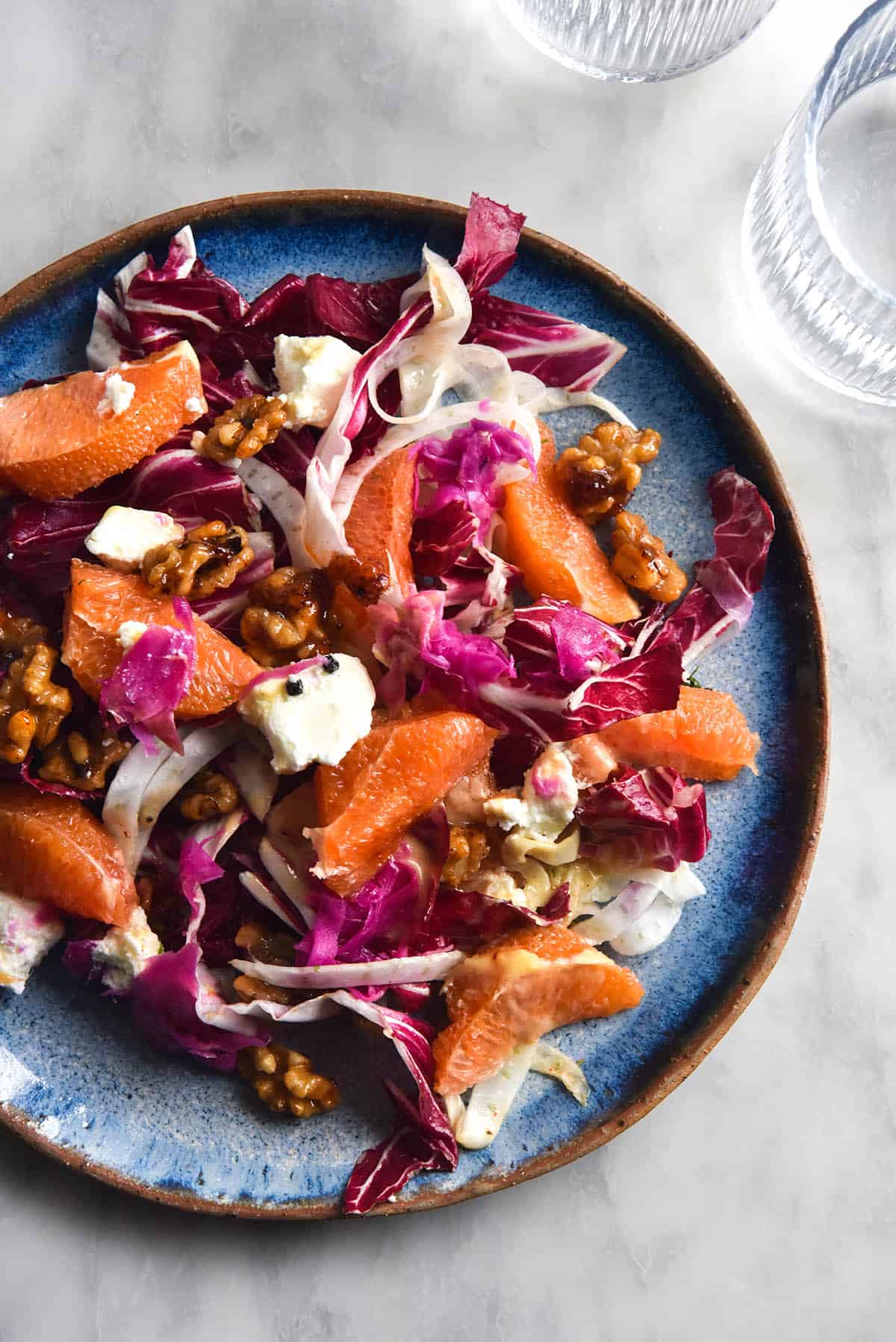 An aerial image of a radicchio, orange, fennel, goat's cheese and chilli maple walnut salad. The salad sits atop a bright sky blue ceramic plate. The scene is set on a white marble table surrounded by textured water glasses that filter gentle sunlight onto the marble 