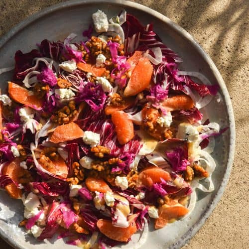 An aerial view of a radicchio orange salad with fennel, goats cheese, chilli maple walnuts and FODMAP friendly faux pickled onion. The salad sits atop a white ceramic serving platter atop a stone backdrop bathed in sunlight. The shadows of a grassy thin leafed plant create a pattern on the stone to the top of the image