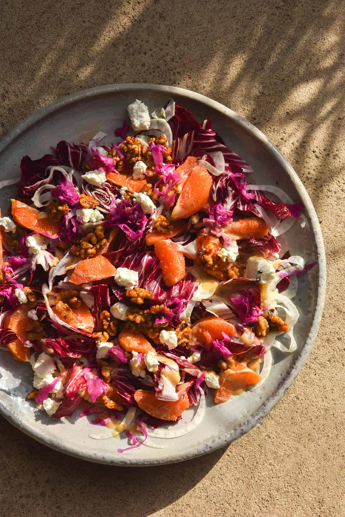 An aerial view of a radicchio orange salad with fennel, goats cheese, chilli maple walnuts and FODMAP friendly faux pickled onion. The salad sits atop a white ceramic serving platter atop a stone backdrop bathed in sunlight. The shadows of a grassy thin leafed plant create a pattern on the stone to the top of the image