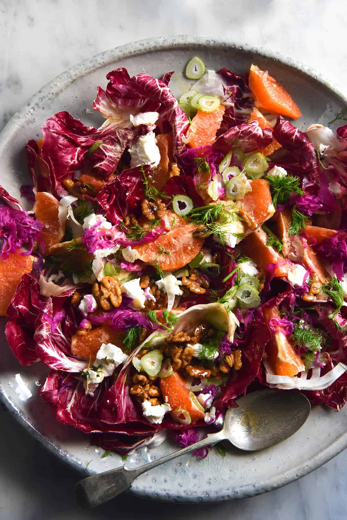 An aerial close up view of a radicchio, orange, fennel, goat's cheese and maple chilli walnut salad. The salad sits atop a large white ceramic plate on a white marble table. A serving spoon sits alongside the salad in the centre bottom of the image