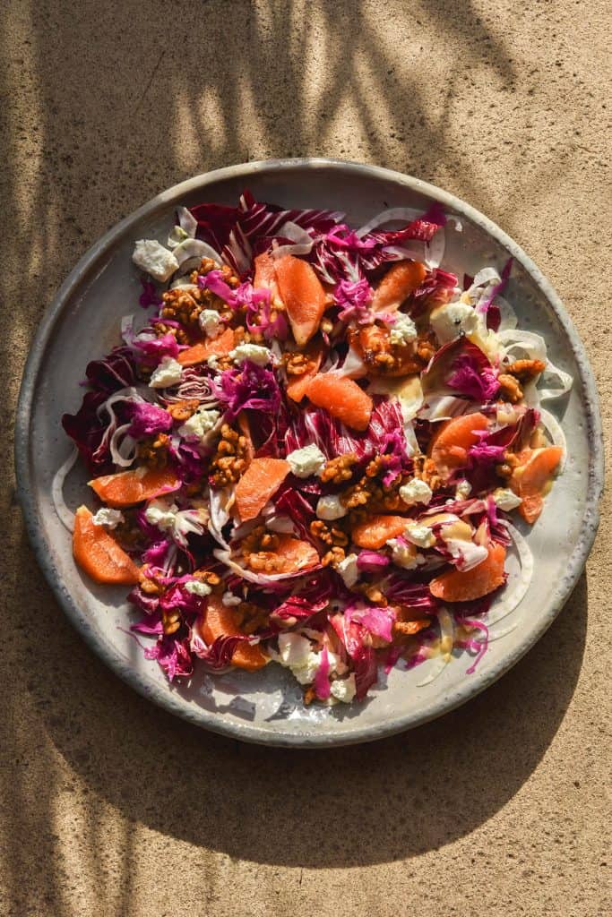 An aerial image of a radicchio, orange, fennel, goats cheese and chilli maple walnut salad on a white ceramic serving platter. The salad sits atop a beige stone backdrop which is bathed in natural light. Thin grassy leaves project their shadow in a pattern across the top of the image and down the left hand side