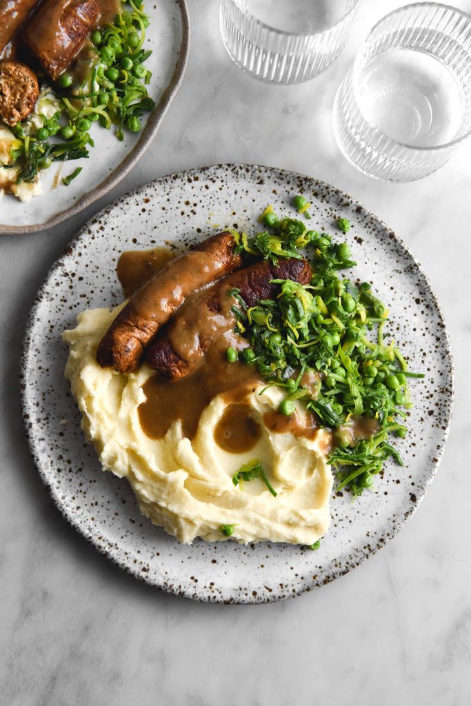 An aerial view of two white plates topped with mash, vegan sausages, FODMAP friendly sauteed leeks and peas and gluten free, vegan gravy. The plates sit atop a white marble table and two sunlit glasses of water sit to the top right of the image