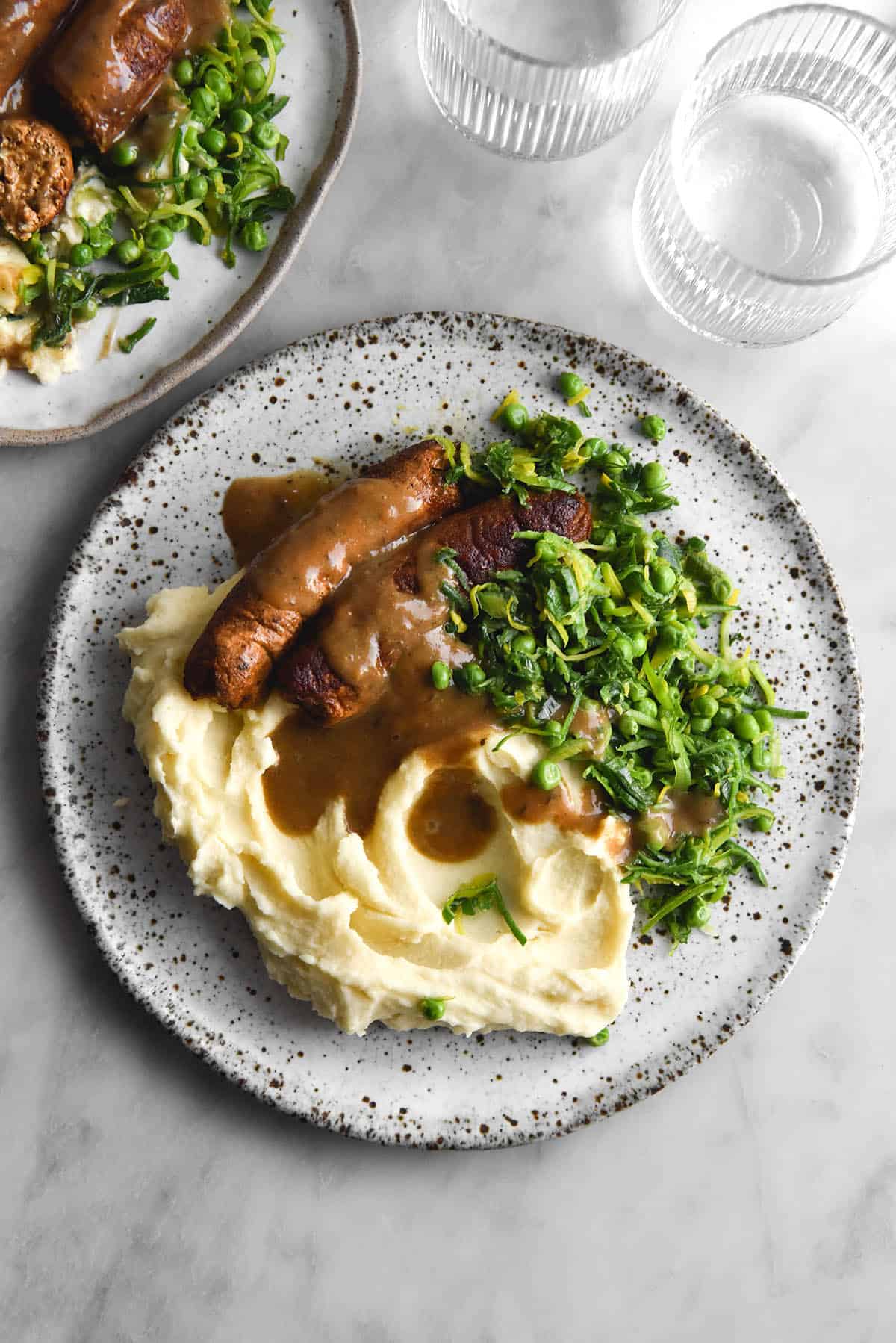 An aerial view of a white speckled ceramic plate topped with mash, vegan sausages, FODMAP friendly leeks and peas and a gluten free vegan gravy. The plate sits atop a white marble table. A second plate of bangers and mash sits to the top left of the image, while two sunlit water glasses sit in the top right hand corner. 