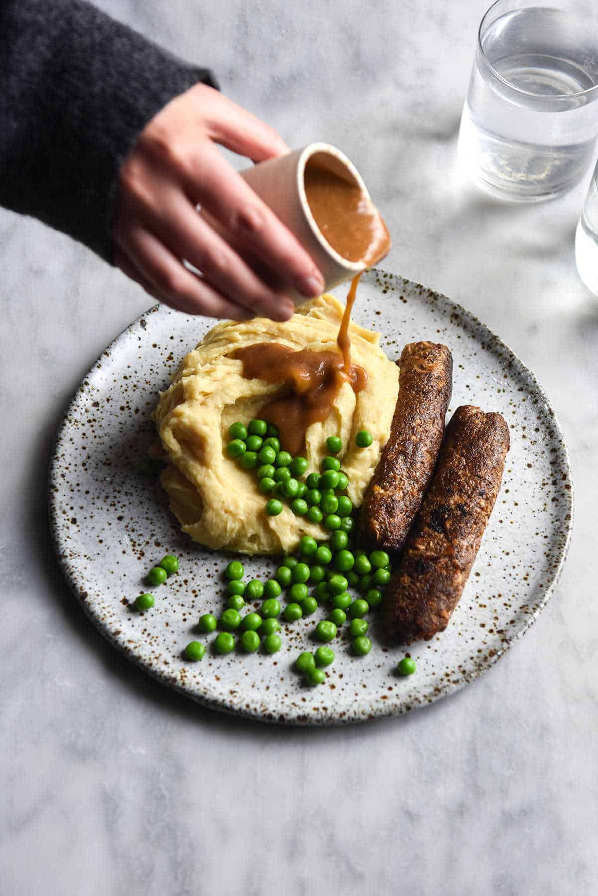 A photo of a white speckled ceramic plate topped with vegan sausages, mash and peas. A hand extends out across the white marble table to pour gravy over the mash potato and some sunlit glasses of water sit to the top right of the image