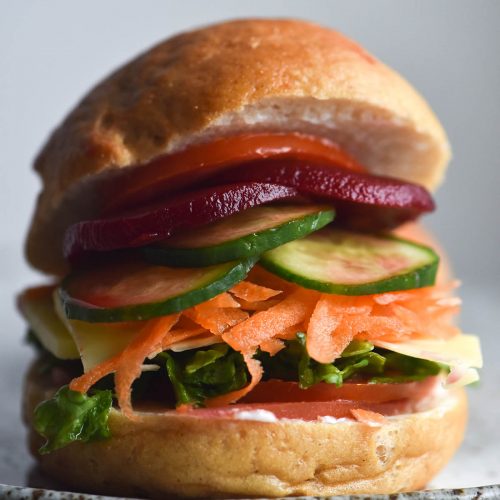 A side on image of a gluten free bread roll filled with salad. It sits atop a white speckled plate against a white backdrop