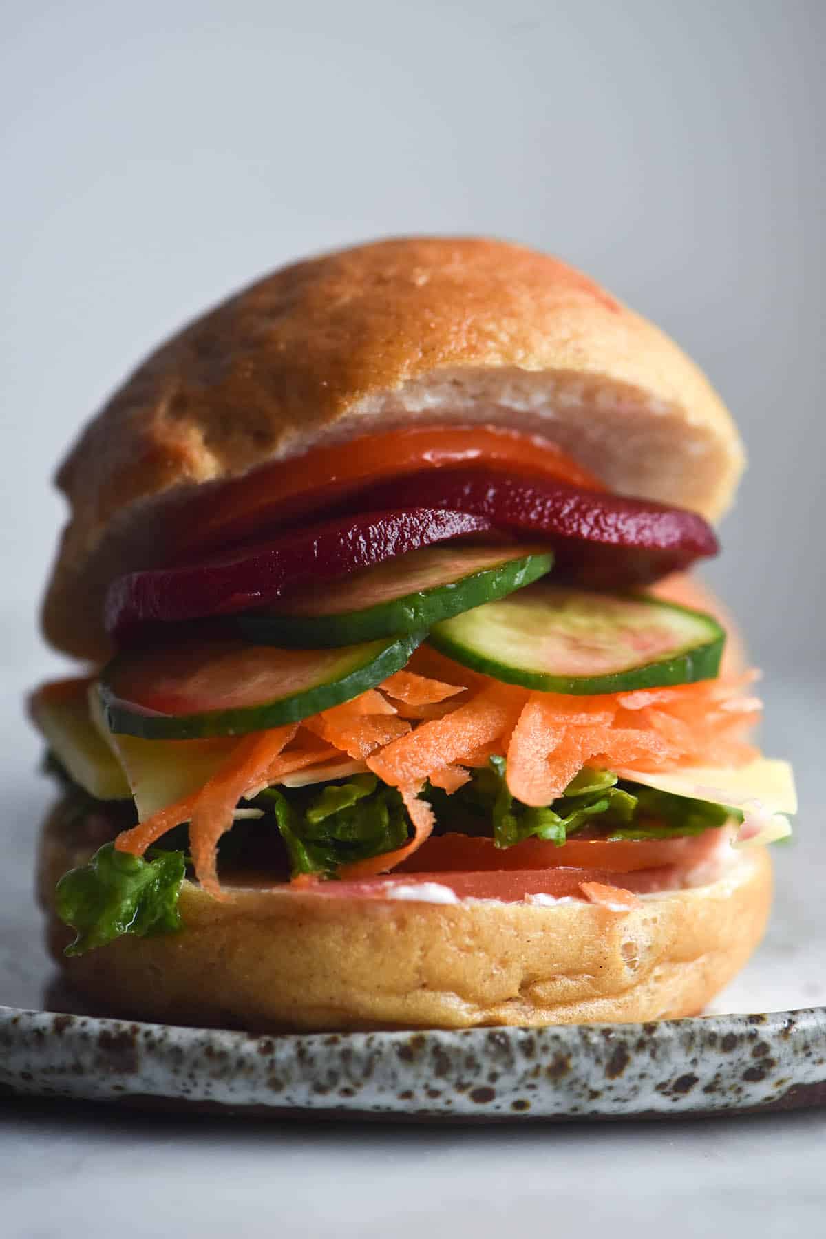 A side on close up view of a gluten free bread roll made into a salad sandwich. The salad sits atop the bun base and the top sits loftily atop the salad. It sits on a white ceramic plate against a white backdrop.