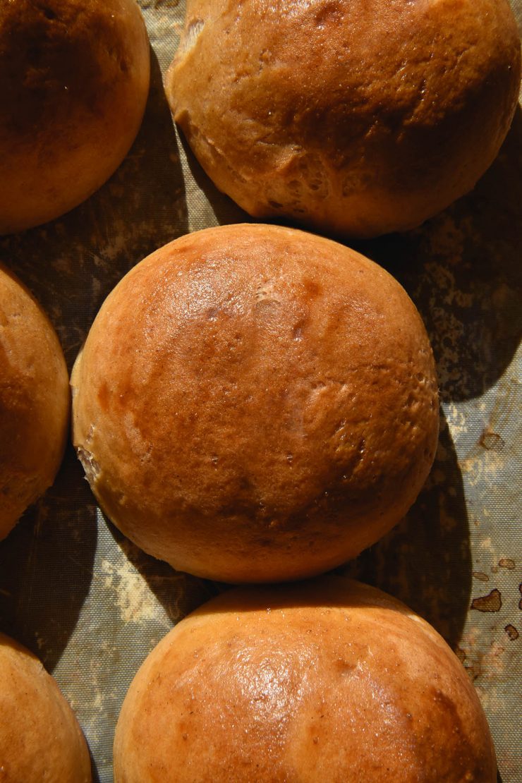 An aerial view of golden brown gluten free bread rolls atop a baking tray. The rolls are bathed in natural sunlight which produces shadows across the image