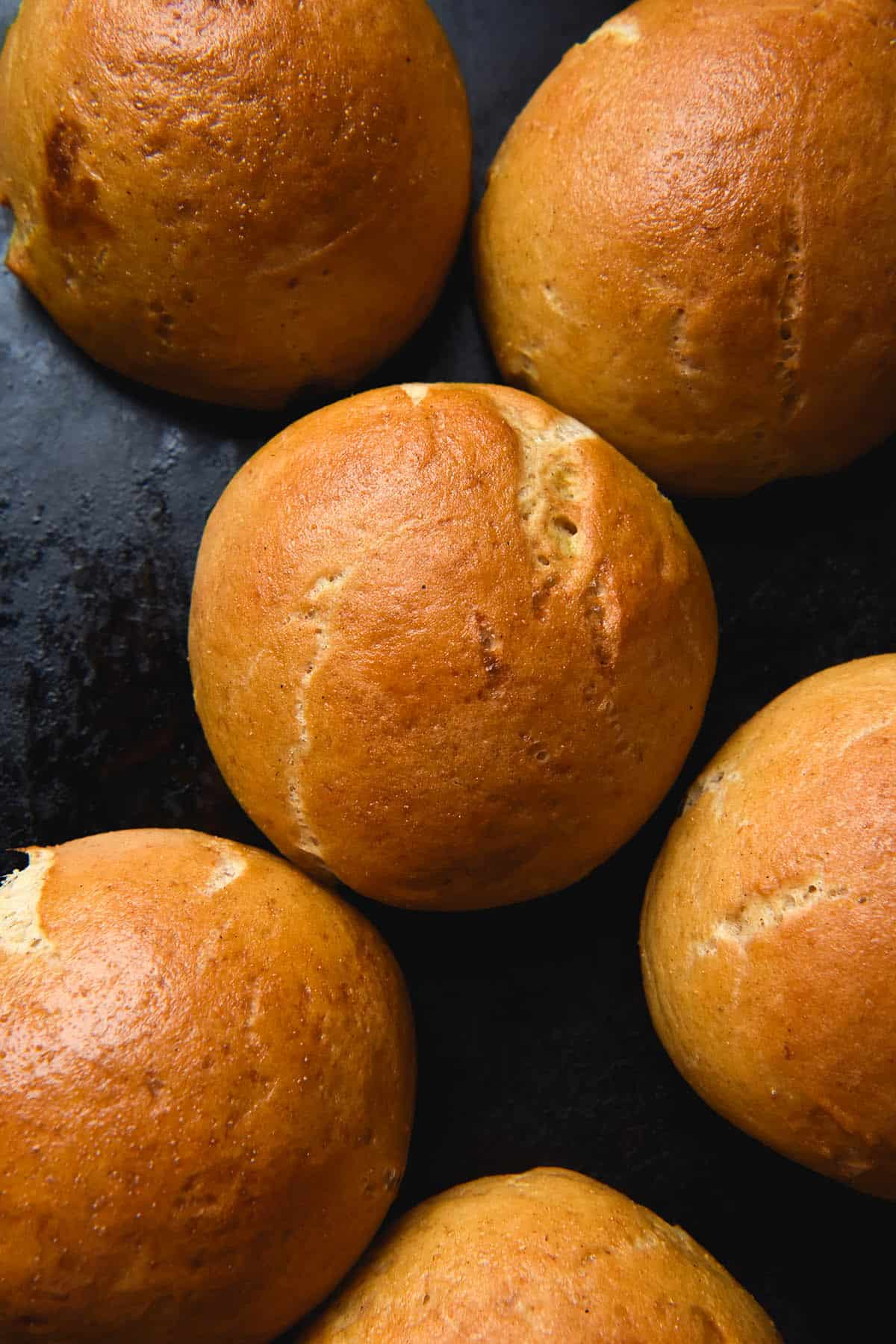 Gluten free bread rolls arranged casually atop a black baking tray. The rolls are perky and golden brown and have cracks from expanding in the oven. 