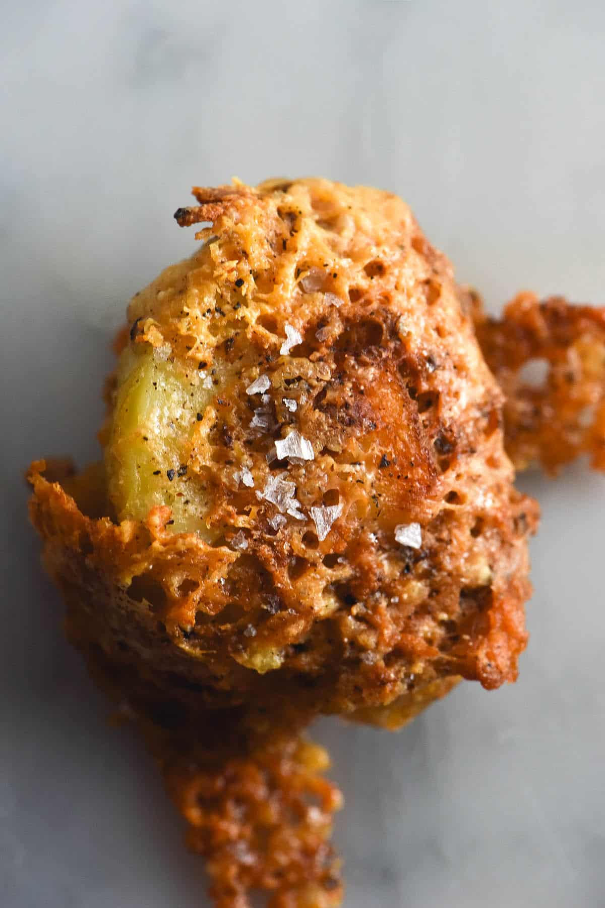 A close up aerial image of a crispy roasted cacio pepe potato. The potato is covered in a crispy parmesan shell flecked with pepper and topped with sea salt flakes. It sits atop a white marble table.