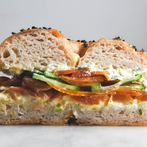 A side on view of a gluten free bagel half, filled with cream cheese, cucumbers and carrot lox. The bagel sits atop a white marble table against a white backdrop