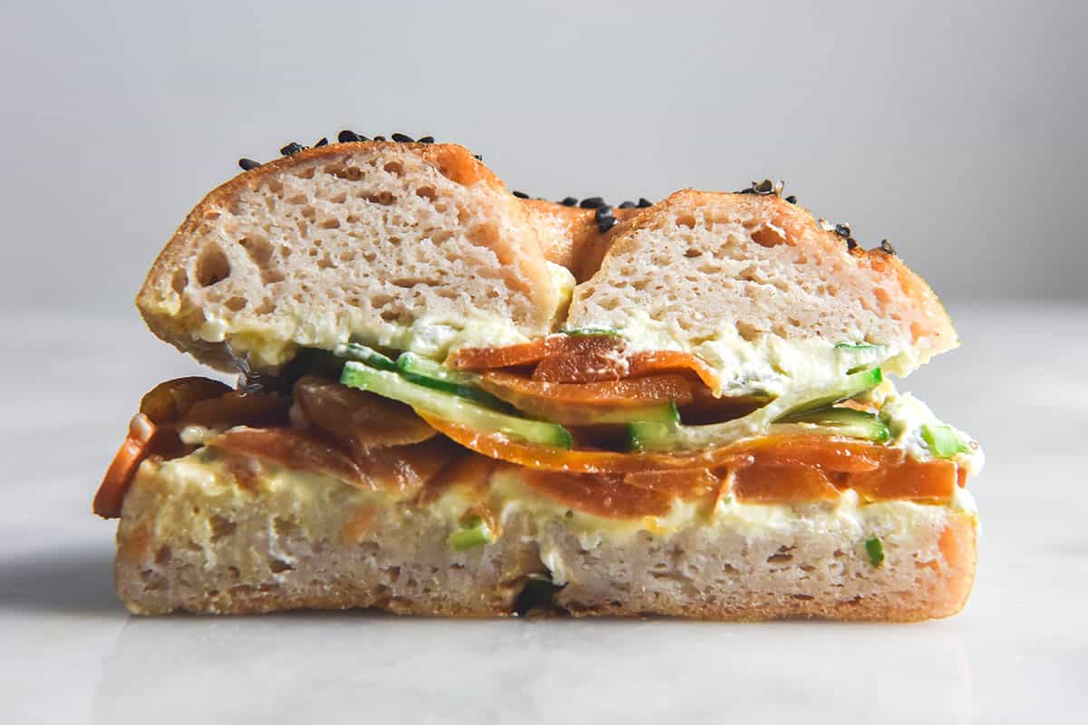 A side on view of a sliced gluten free bagel half filled with cream cheese, cucumber and easy carrot lox. The bagel sits atop a white marble table against a white backdrop