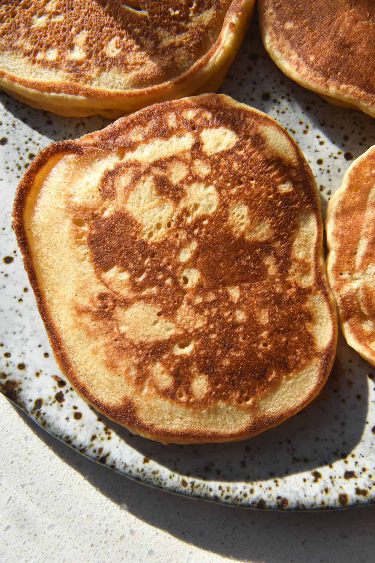 An aerial close up image of cassava flour pancakes atop a white speckled ceramic plate. The pancakes are bathed in bright sunlight and have golden brown exteriors. 