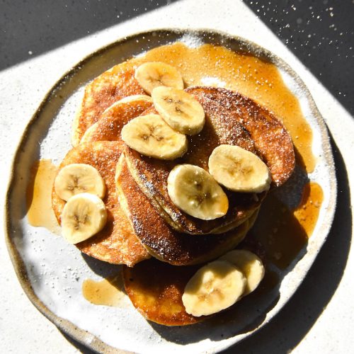 An aerial view of cassava flour pancakes topped with banana coins, maple syrup and a dusting of icing sugar that rains down from the top of the image. The pancakes sit atop a white marble plate against a white benchtop which is drenched in sunlight with deep contrasting shadows around the corner of the image