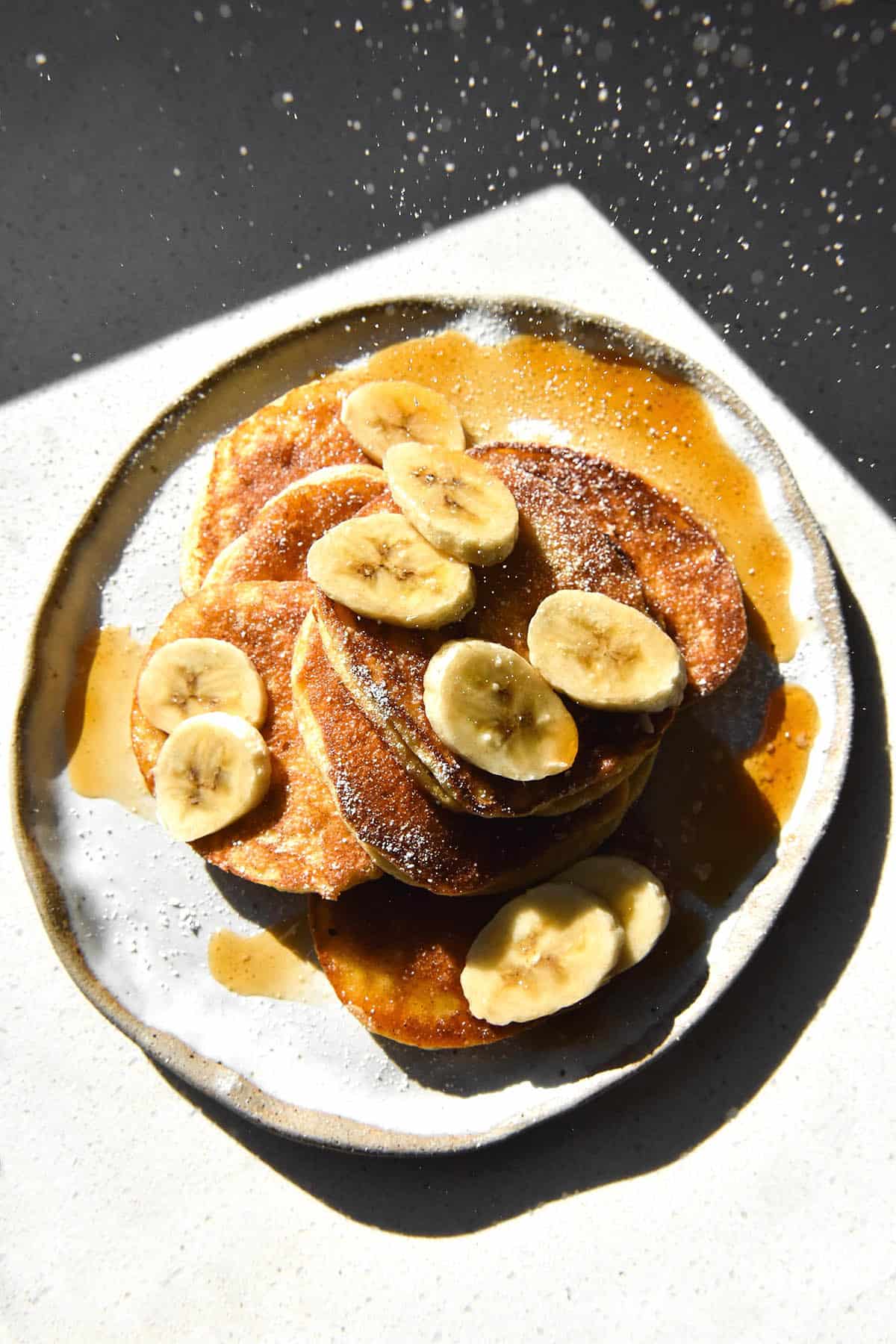 An aerial view of cassava flour pancakes topped with banana coins, maple syrup and a dusting of icing sugar that rains down from the top of the image. The pancakes sit atop a white marble plate against a white benchtop which is drenched in sunlight with deep contrasting shadows around the corner of the image