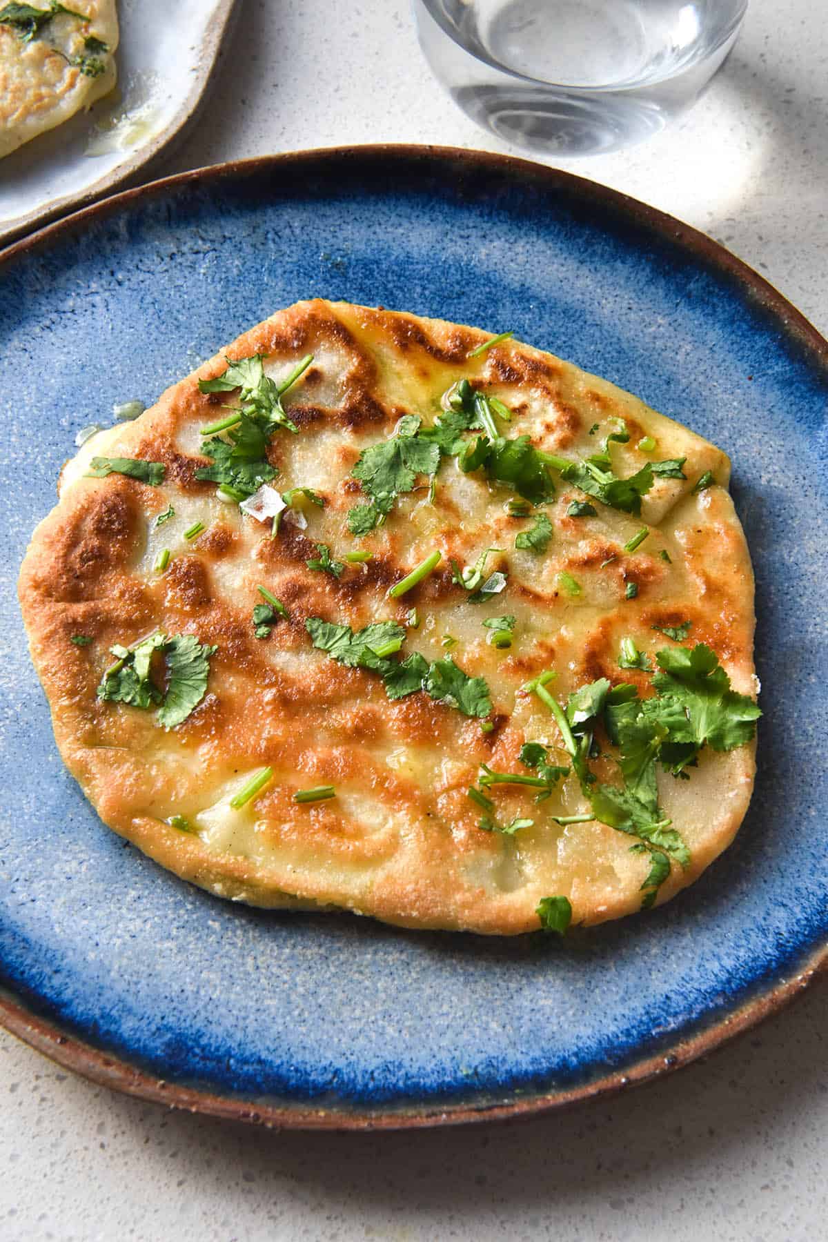 A close up image of a gluten free cheesy naan topped with garlic infused ghee, chopped parsley and sea salt flakes. The naan sits atop a sky blue ceramic plate on a white benchtop. Another plate of naan and a water glass sit to the top of the image.