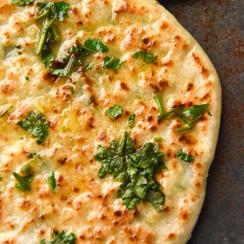 An aerial, close up view of a gluten free cheesy naan topped with garlic infused ghee, chopped coriander and sea salt flakes. A second naan sits in the top right of the image, which is set on a blue steel backdrop